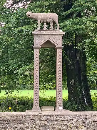 The figures of Romulus and Remus are shown suckling the she‑wolf. The figures are set on a rectangular plinth that is carried on four square columns. The whole stands on a pedestal at the rear edge of a stone boundary wall. Beech trees, without leaf, are shown to the right of the image.