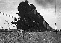 A black and white photo of a large steam locomotive hauling cargo taken from a low angle on a cloudy day.