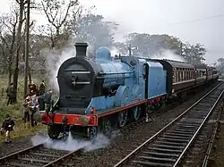 A blue steam locomotive stopped with a number of brown passenger coaches behind her. A small crowd stands beside the track behind her, while a small jet of steam erupts from between her wheels.