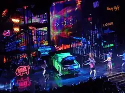 Bird's eye view of a stage, showing large scaffoldings, neon signs and a green car lying in the middle.