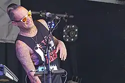 smiling at the crowd a man with a tidy black mohawk and mirrored sunglasses is behind a microphone on stage