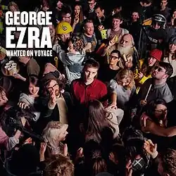 George Ezra is looking towards the camera, standing in the centre of a crowded room. His name and the album title are in the top corner, in large white lettering.