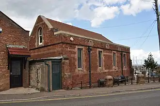 Rectangular church like building in red stone with contrasting natural light stone features and high windows against a light sky