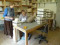 Two women are in a workshop full of moulds. The woman on the left, Griselda Hill, is standing, while the other woman sits with an unpainted pig in front of her.