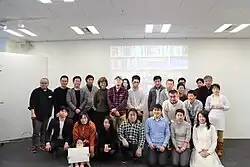 A group of Japanese Wikipedians standing in front of a wall. Kitamura, who is holding a book, is standing at the rightmost place on the second row from the bottom.