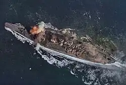 A close view of a large warship from almost directly overhead. Her wake is streaming out behind her and two trails of smoke are visible: a faint plume near her smokestack and a much thicker white plume partially obscuring her foremost main gun turret.