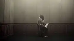 The image shows a man in a dark room, sitting at a chair. He has long hair covering his face, and a white wreath of flowers around his neck. He is holding a white picture book.