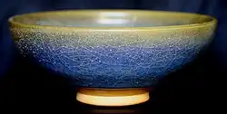A ceramic bowl is viewed from one side. The glaze has a crazed and mottled finish, varying from yellow at the rim of the bowl to blue below.