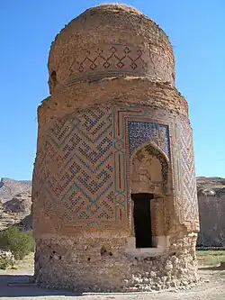 Stubby cylindrical structure with faded and partly ruined patterns, topped with an ornate dome. Yellow flowers in foreground and steep cliffs with grass among the crags in the background.