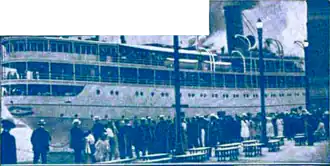 Blue-tinted photograph of a ship, with many people standing in the foreground.