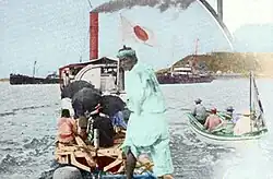 In this picture, a small steam boat is towing a raft and some rowboats toward shore.