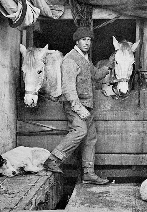 Photo of a man standing in front of two narrow horse stalls, with a sleeping dog behind him