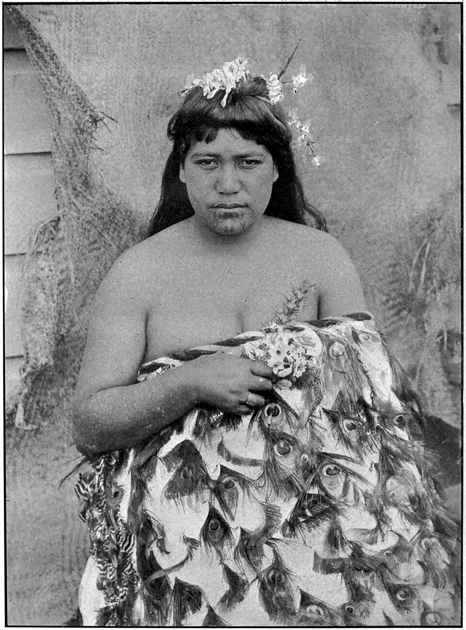 A Māori woman with facial moko, flowers in her hair, and wearing a traditional cloak.