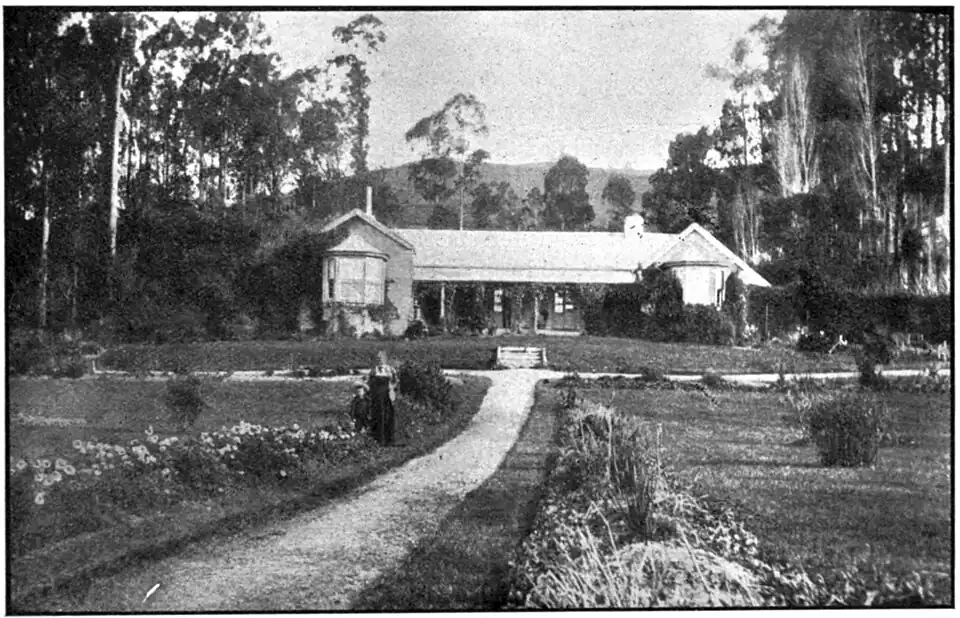House with bay windows at each end, gardens and paths in front with trees to either side and behind, and a lady and child standing beside the main path