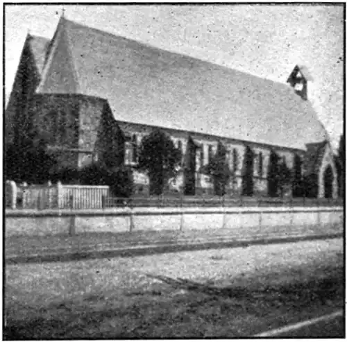 Stone church with bell tower and entrance at one end (on the right) and a low wall, path and road in the foreground