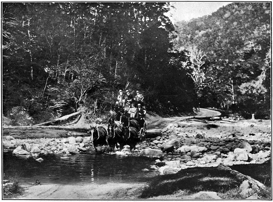 Coach crossing a river with a road and trees in the background