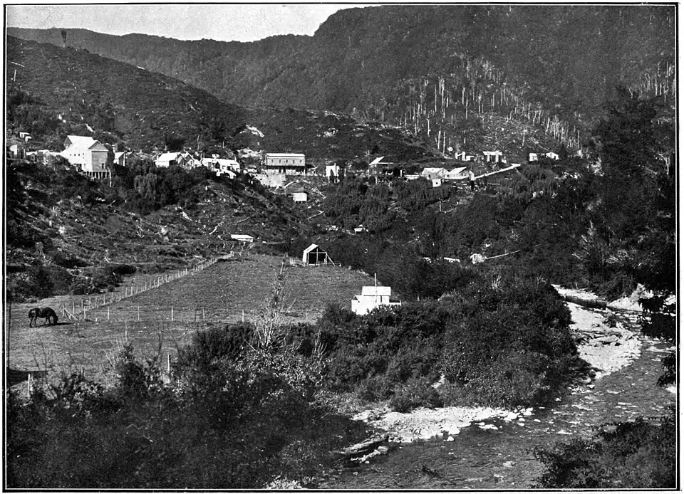 The gold-mining settlement of Lyell with houses and buildings above a cleared slope with tree stumps and a paddock, a small river in the foreground and bush covered hills behind