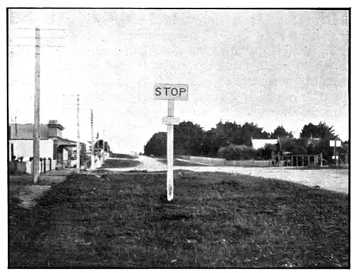 Stop sign next to a dirt road with houses and buildings on each side, and power poles on the left-hand side