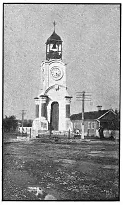 An elaborate clock tower with two clock faces visible and a covered bell tower with finial above it, in the middle of a dirt road with power lines and a house on the right side