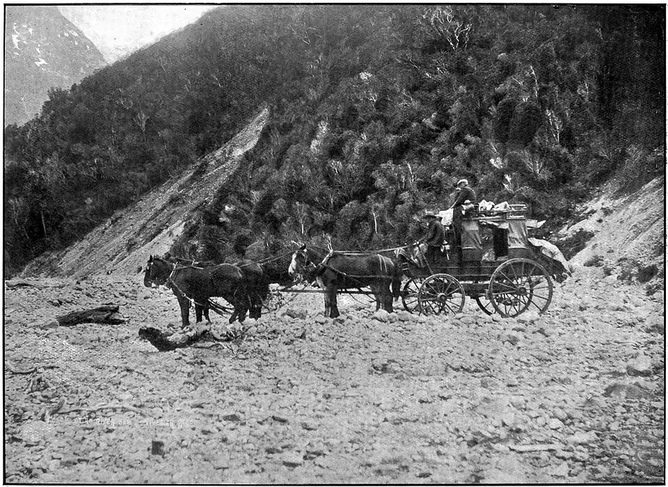 A group of people sitting on a horse-carriage. Behind them is a mountain covered in bush, there is another in the distance.