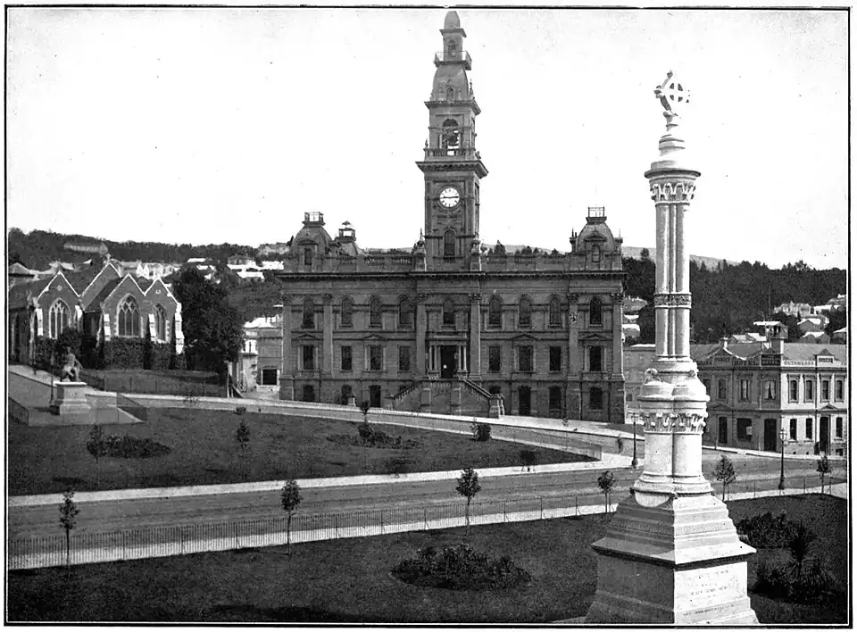 The city centre of Dunedin. On the left of the photo is a church, the size of which is dwarfed by the larger building in the middle of the photo, which has a clock face on the tower. There is a statue in the foreground of the photo that has a cross inside a circle at the top.
