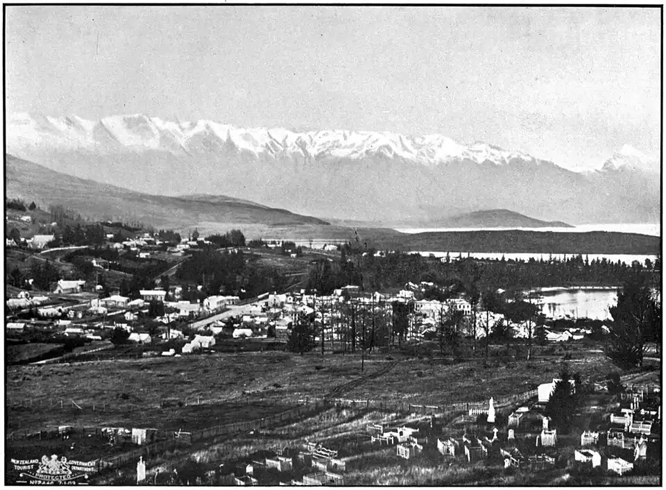 Houses of a small settlement at the edge of a lake with a range of tall snowy mountains behind.