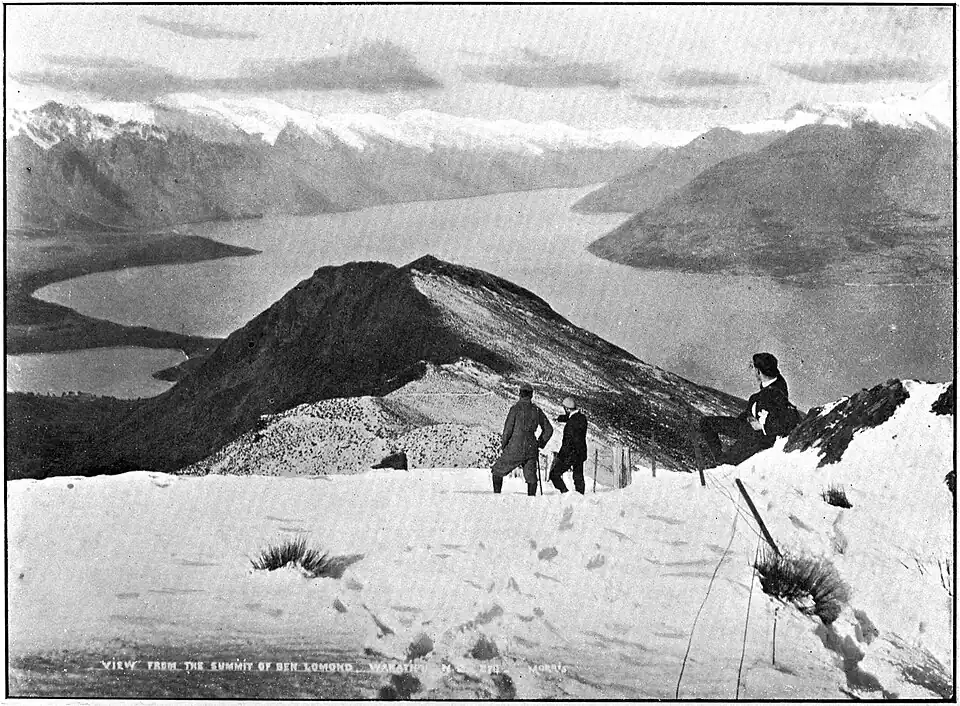 Three climbers in tweedy outdoor clothes on top of a snowy mountain, looking down at a large lake with more snowy mountains in the distance.