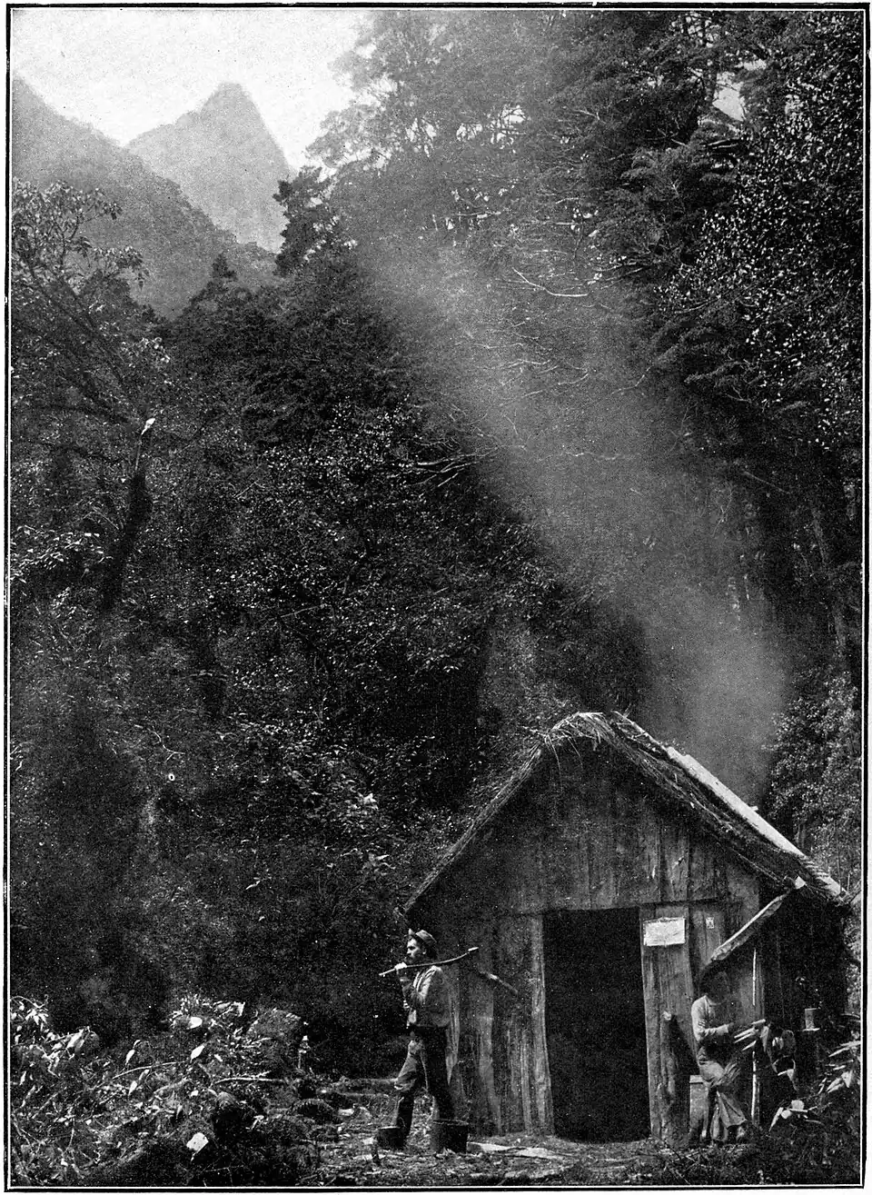 A person walking out of a hut, they are holding an axe. There is another person sitting outside the hut.