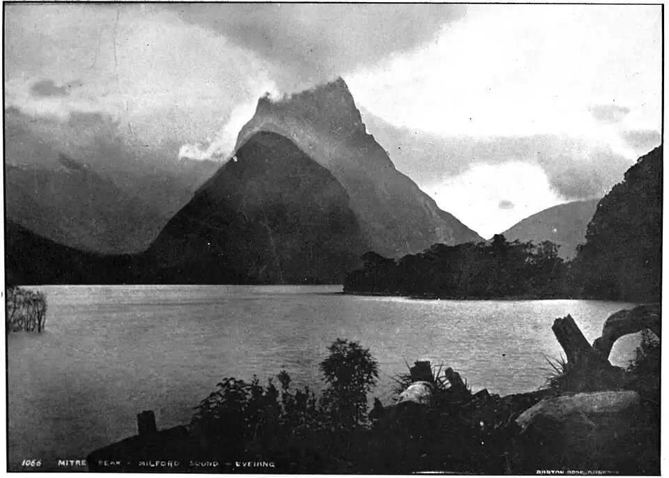 Photo overlooking a lake, there are trees at the bottom of the photo while there is a body of land and mountains in the distance. The scene is very foggy.