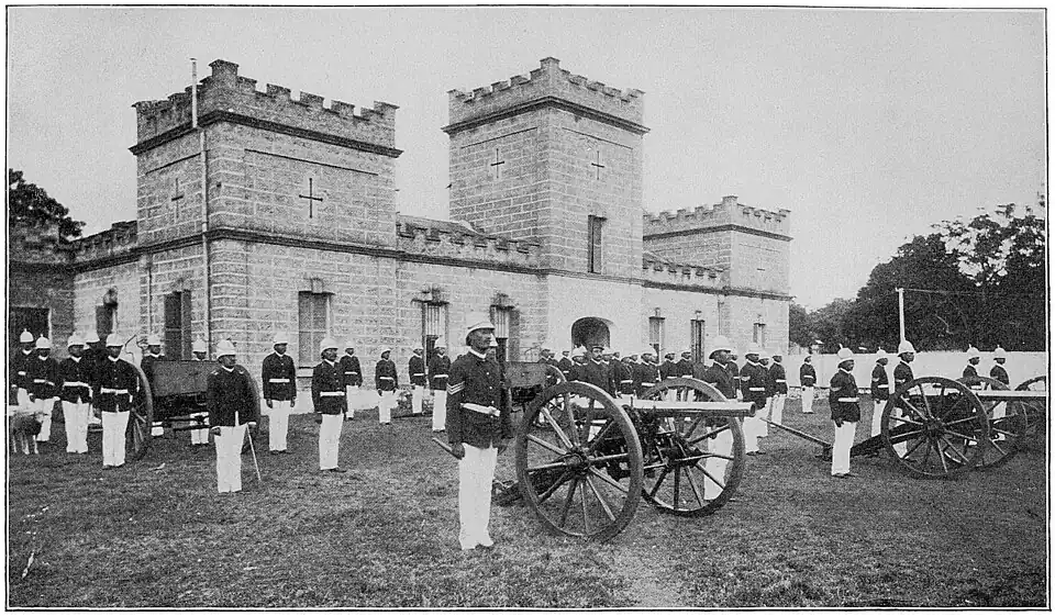 Several soldiers standing along with cannons in front of a brick building with turrets