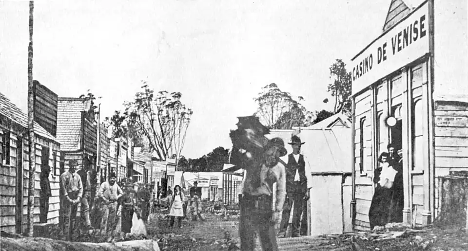 Napoleon Hill, Ahaura, Westland. A typical mining town of the mid sixties. Street scene with wooden buildings on either side, some trees at the far end, men milling about. Note the Casino De Venise, with dancing girls in doorway.