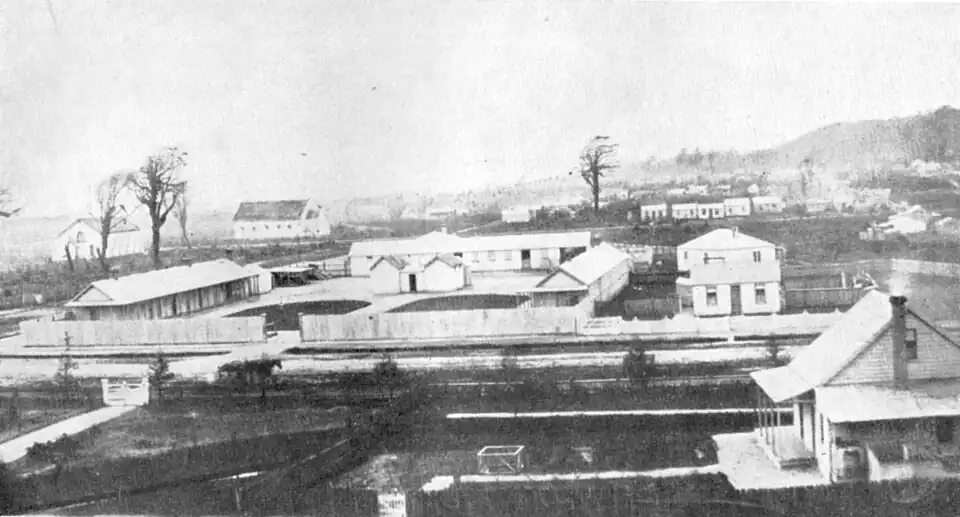Several large buildings with two lone trees. In the distance to the right is a hill.