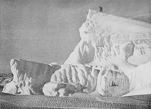 Photo of the jumbled edge of a tabular iceberg with the small figure of a man at its peak