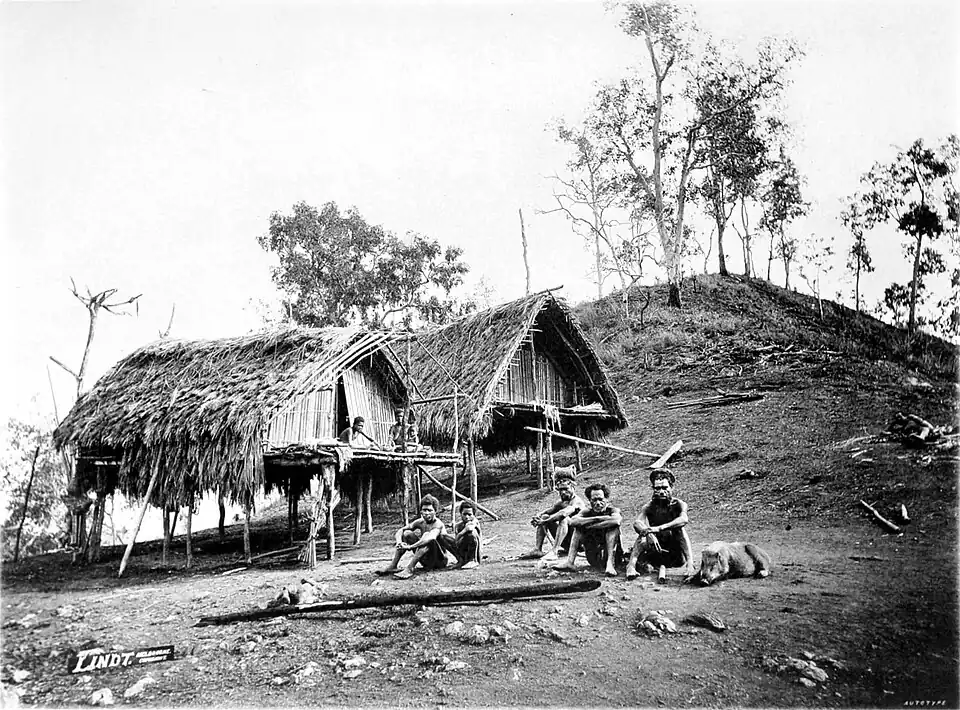 Black and white photograph of a group of crouching men and a pig, in front of two huts.