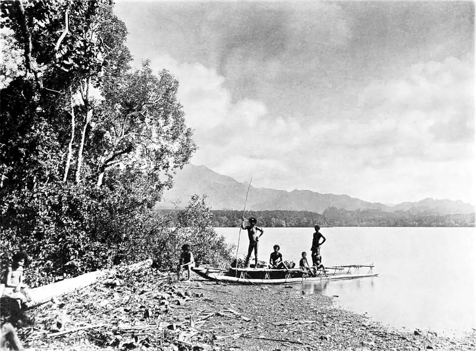 Black and white photograph of three men and two children with a canoe moored on a beach. Trees and driftwood line the beach. In the background, across the water, more land and mountains can be seen.