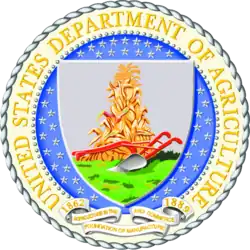 The Seal of the United States Department of Agriculture - the name of the department in a circle around a shield, which shows a plough and haystack on a gray field, with stylised grass or plants in the bottom third.
