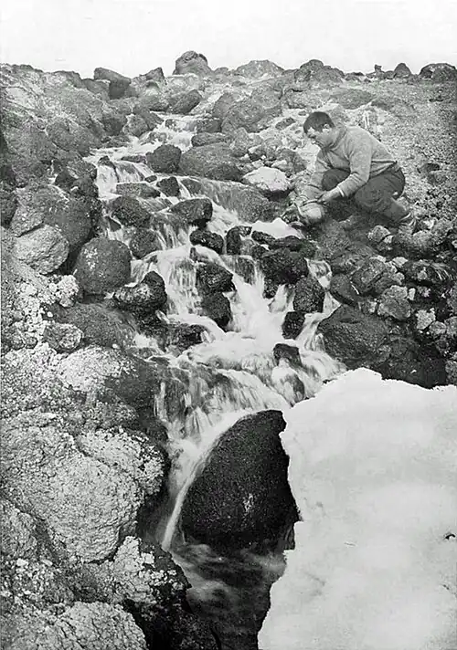 Photo of a man with a kettle crouching by a cascade running down a rocky slope