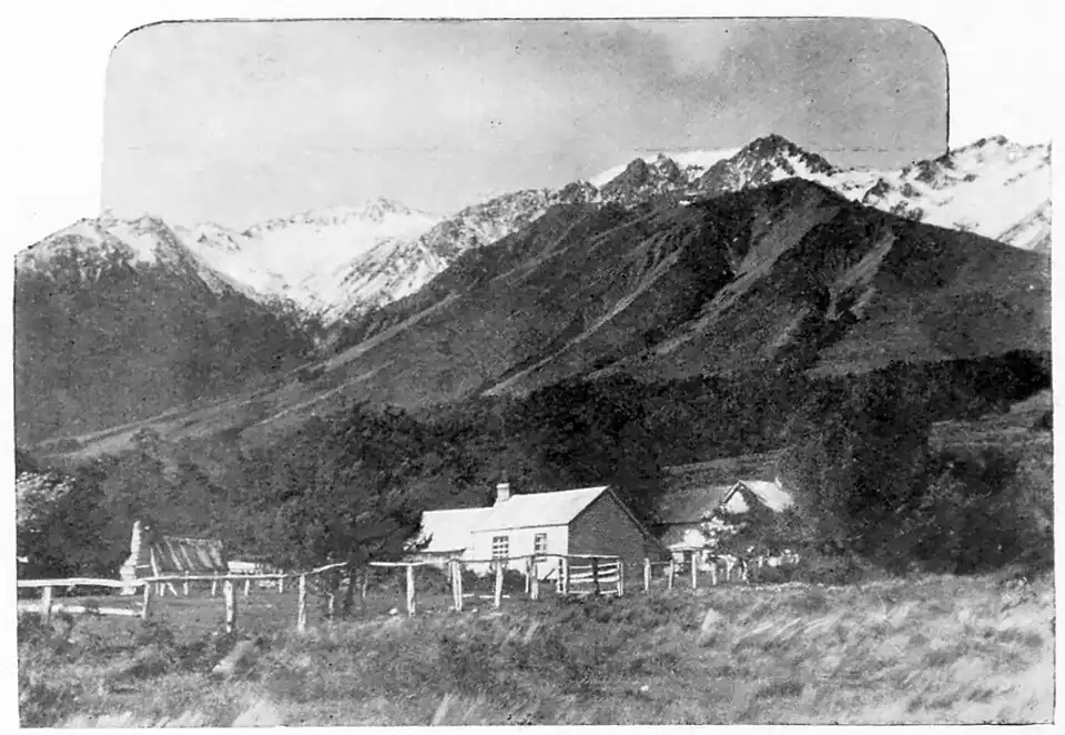A homestead and other buildings surrounded by a fence, with trees and bush behind them, at the bottom of a mountainous slope with a snow-capped mountain range behind.