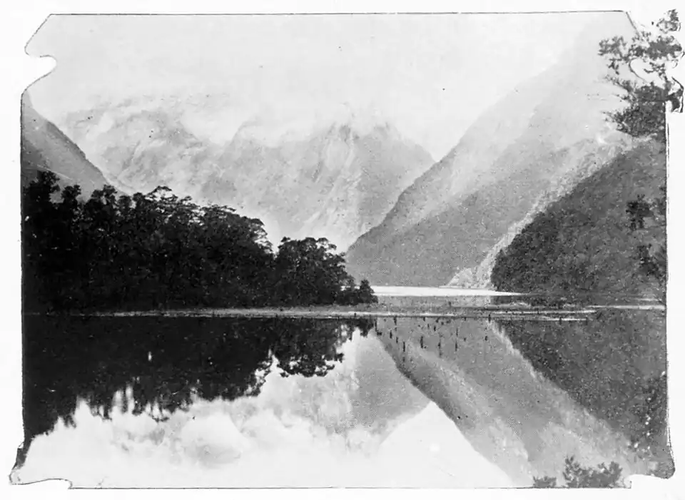 Snow covered mountain range in the background with bush and trees at the base and a lake in the foreground.
