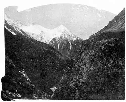 Snow-capped mountain range in the background, with a gorge in the centre with rocky, barren steep sloping hills on either side.