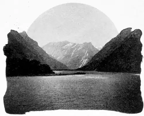 A calm, flat lake with hills on either side in a V-shape about the center of the image, and mountains in the background.