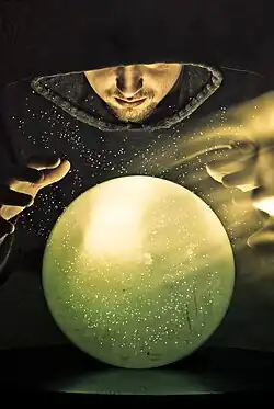 Colour photograph of a hooded wizard gazing into a green crystal ball with the ghost image of his face floating to the right.