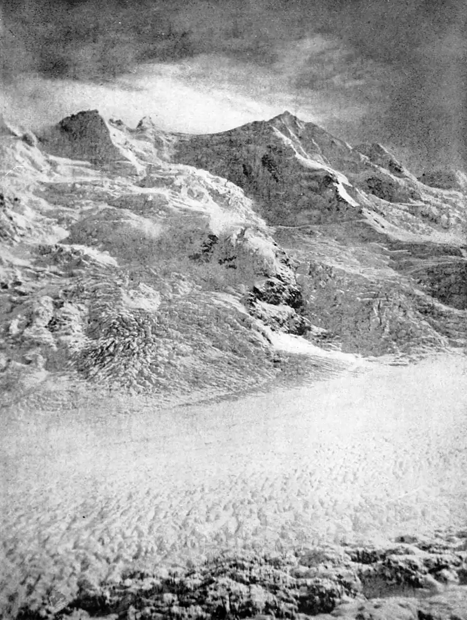 Glacier in the foreground with snow covered mountains and scattered clouds in the background, with a scattering of exposed rocks
