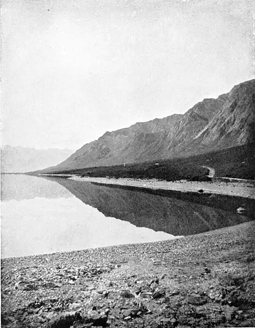 A rocky hill on the right, with trees on a flatter section at the base. A track goes through the trees to the rocky shore of a lake. The lake has a mirror image of the hills reflected in it.