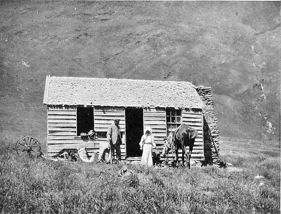 A basic hut sitting in an open grassy plain with a steep bare hill behind it. The hut has a gently sloping roof, a doorway in the middle with a window on either side (only one with some glass), and a chimney at one end. Two people and a horse are in front of the hut.