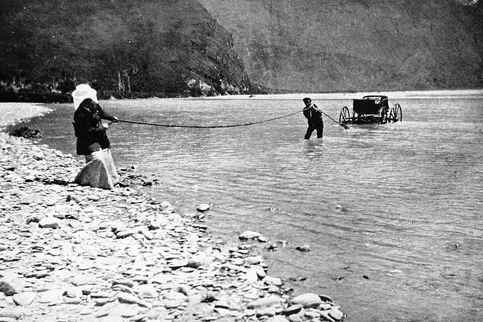 A four-wheeled carriage in a calm river is pulled ashore with a rope by two men - one near the carriage with the rope over his shoulder, and the other pulling the rope from the gravel river bank.