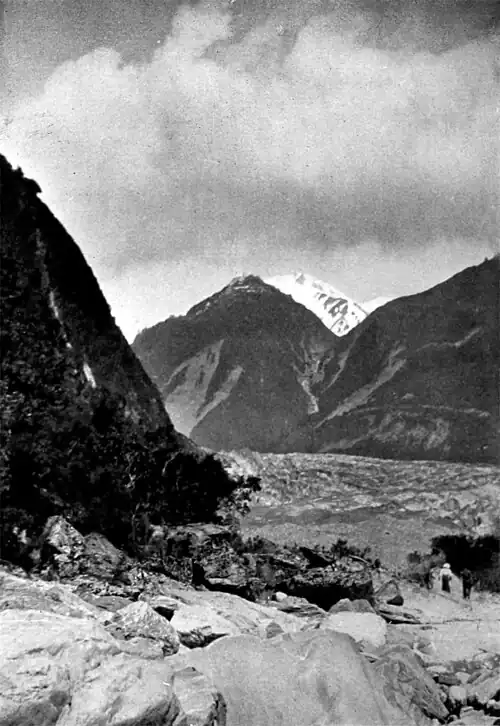 Rocky foreground with a hut, then the start of the Fox Glacier with steep hills on either side, and snow-capped mountains in the background with a cloudy sky