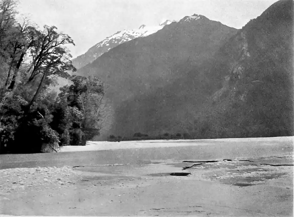 There is a snow-capped mountain in the distance, before this there is a steep bush clad hill that comes down to thea river's edge, some trees to the left, with a beach in the foreground