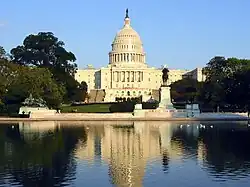 Color photograph of the United States Capitol building, a neo-classical structure framed by trees on each side and water in the foreground
