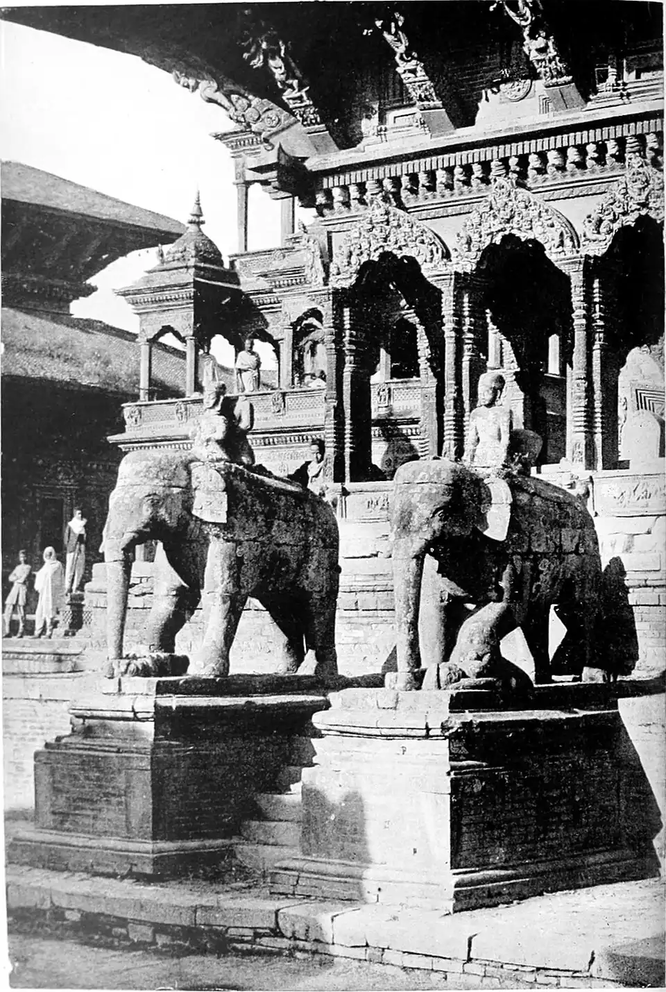 Black and white photograph of two statues of mounted elephants on plinths either side of a narrow set of steps. A building with ornate arches and columns fills the background.